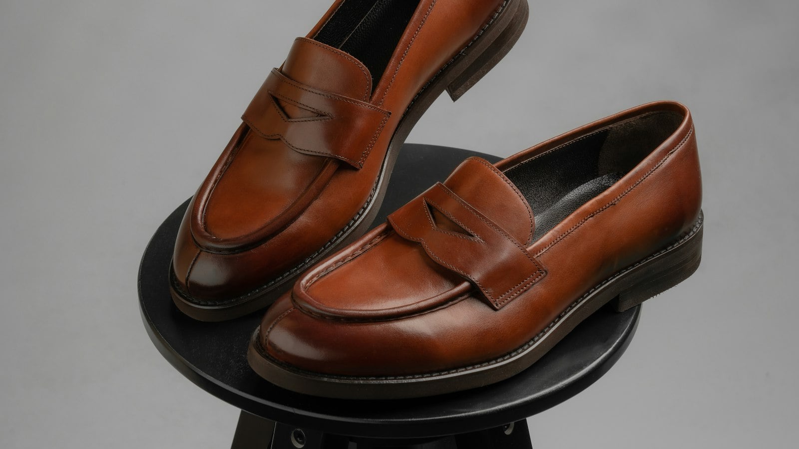 A pair of brown leather penny loafers balanced on a dark stool against a grey background (photo: Husien Bisky / Unsplash)