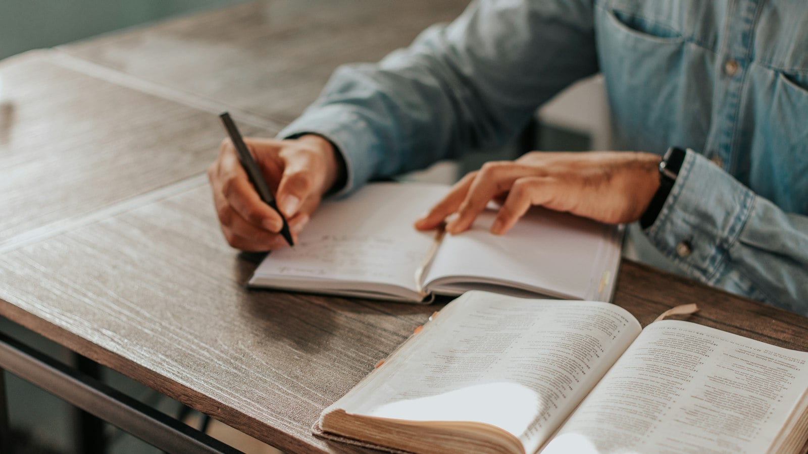 Hands writing notes in a notebook beside an open textbook on a wooden table (photo: Fa Barboza / Unsplash)
