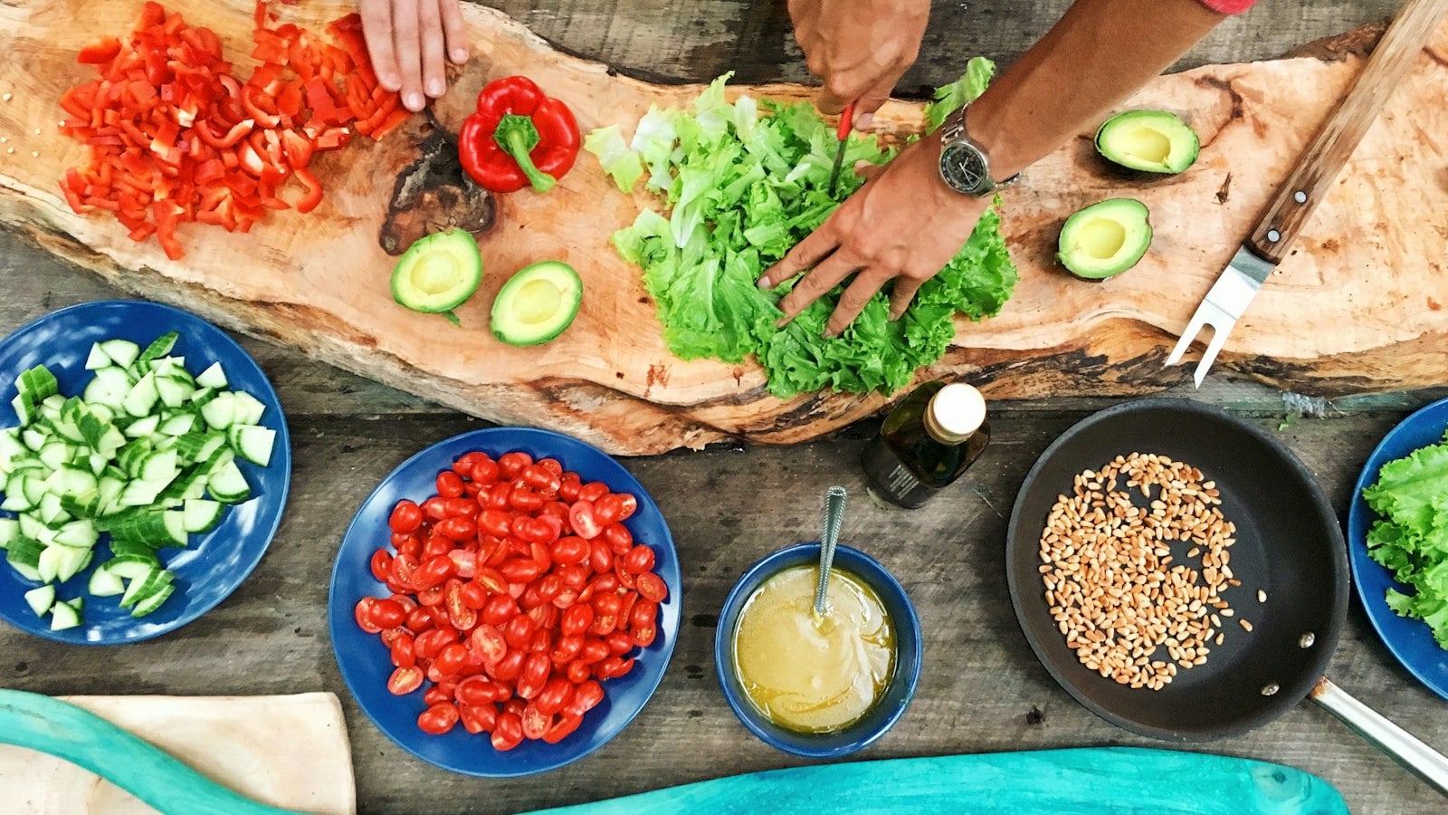 Hands preparing vegetables on a wooden board beside bowls of tomatoes and cucumber (photo: Maarten van den Heuvel / Unsplash)