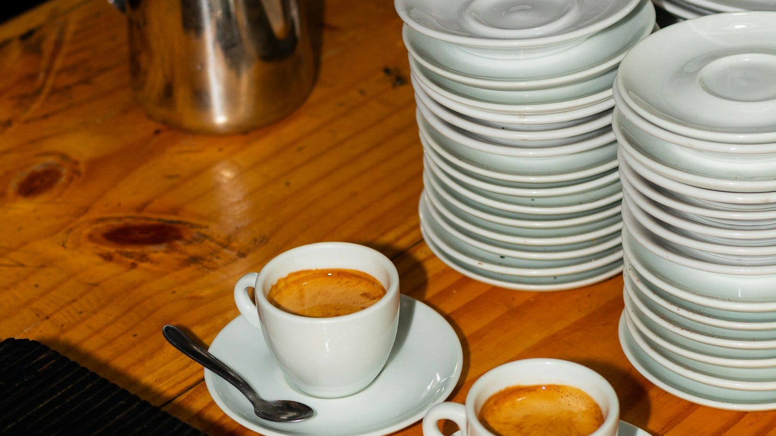 Two espressos in white cups beside tall stacks of white saucers on a wooden café counter (photo: Romario Roges / Unsplash)