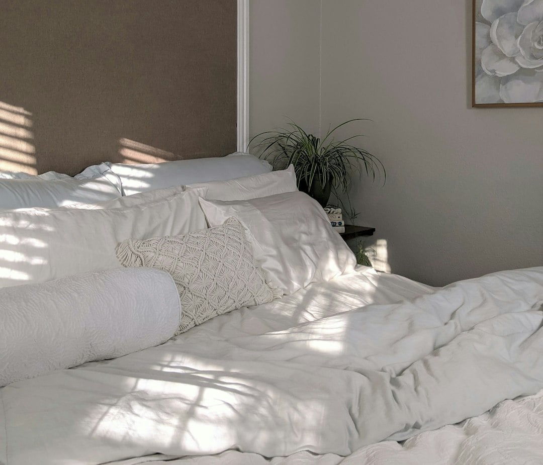 A bed heaped with white bedding and pillows beside an upholstered headboard and houseplant