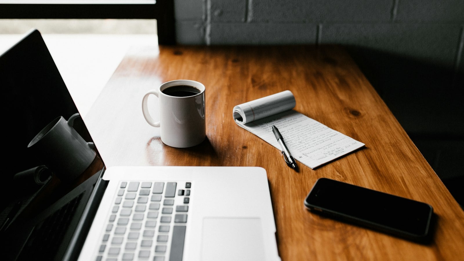 A laptop, a cup of coffee, a notepad and a phone on a wooden desk (photo: Andrew Neel / Unsplash)