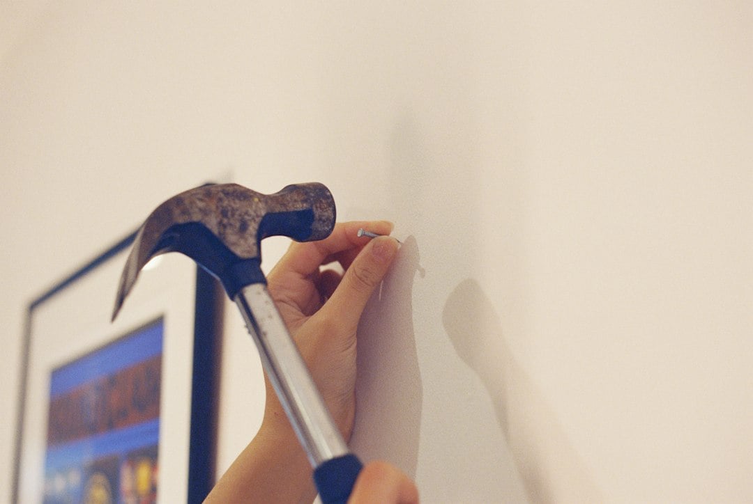 Hands position a nail against a white wall while holding a hammer, with a picture frame waiting below