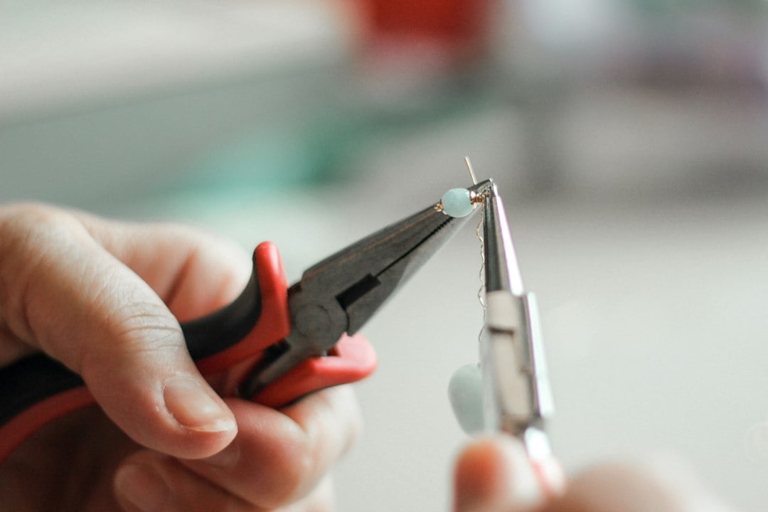 Two hands use round-nose and flat-nose pliers to close a wire loop on a jewelry finding.