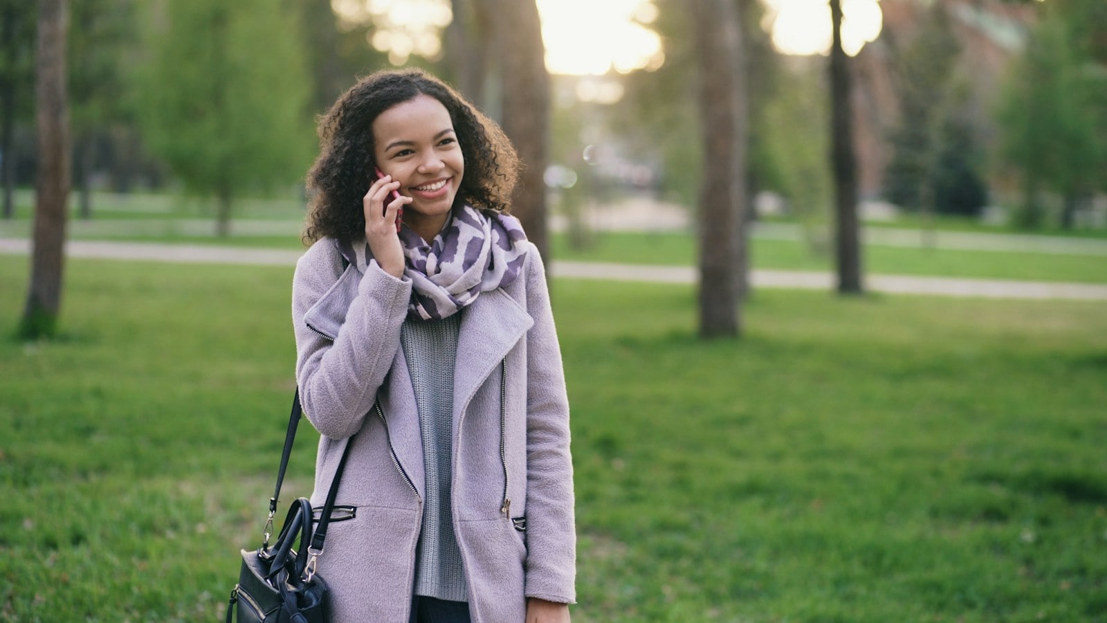 A smiling woman in a lilac coat and scarf talking on her phone in a green park (photo: Vitaly Gariev / Unsplash)
